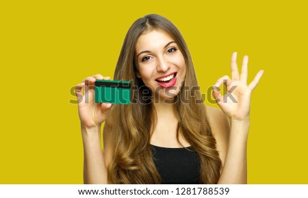 Smiling young woman showing credit card and showing OK sign over yellow background