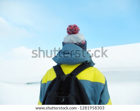 A boy in a colorful jacket, a cap with a bubo and a backpack on his shoulders against the backdrop of a snowy mountain and a winter blue sky. Back view