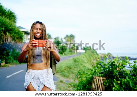 Positive smiling african woman taking pictures of nature environment using camera and application on smartphone device during time for discovering world, happy hipster girl enjoying summer holidays