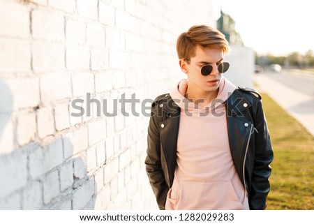 Handsome fashionable young hipster man in black sunglasses in stylish black leather jacket in a pink sweatshirt standing near a white brick wall against the background of the sun rays. Attractive guy.