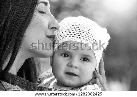 Mother kissing and holding small daughter baby girl cute hazel-eyed kid tiny little child wearing white flower beanie hat outdoor sun spot summer day on blurred green background, horizontal picture 