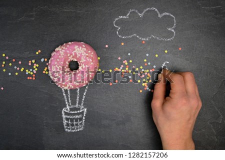 On a dark stone surface a basket of a hot air balloon was drawn, a pink donut with white sprinkles forms the balloon concept with donut and chalk