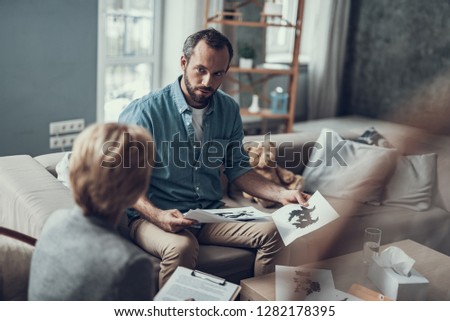 Serious middle aged man sitting on the sofa and looking attentively at his psychotherapist while holding pictures from test