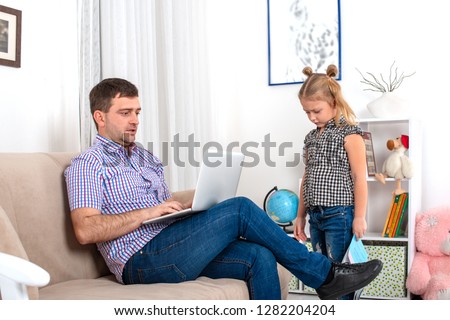 A girl with a textbook went to her dad and asked him to help with the lessons.  Dad is sitting on the couch with a laptop, he is very busy.  The daughter is ignored by the father, she is upset 