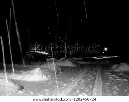 a country house by the road, surrounded by snow drifts, in the dark, at the end of the street a lamppost illuminating the road. In the open air, with precipitation - snow falls. in black and white.