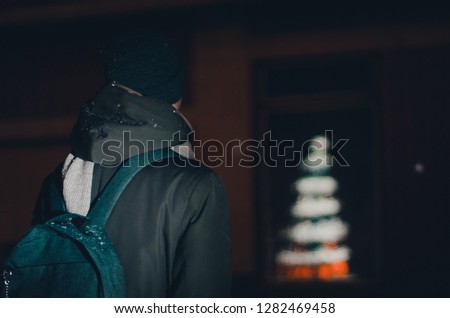 A man in winter clothes and a backpack outside in winter season. The shot is made from the back view. Bokeh lights of decorated Christmas tree on the blurry background of Christmas market street.