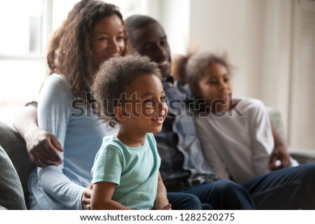 Happy black family with children sitting on couch watching tv together, african american parents embrace kids relaxing on sofa laughing enjoying funny cartoons or movie having fun on weekend at home