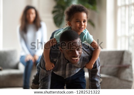 Happy toddler boy playing with father at home, black dad giving cute mixed race child piggyback ride carrying little son on back, african american daddy and kid having fun spending time together