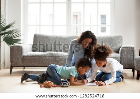 Loving black mom and little mixed race children drawing with colored pencils in living room, happy single african mum helping kids with creative activity at home, underfloor heating, family hobbies