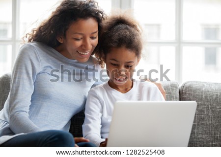 Smiling african american mother and kid daughter have fun shopping online at home, happy black mom teaching little mixed race child girl learn use laptop, watch cartoon, make video call on computer