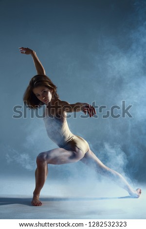 Dancing in cloud concept. Muscle brunette beauty female girl adult woman dancer athlete in dust / fog. Girl wearing dance clothing making dance element performance on isolated grey / black background