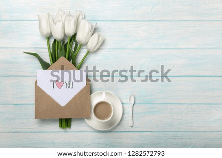 A bouquet of white tulips and a cup of coffee with a love note on blue wooden boards. Top view with place for your congratulations.