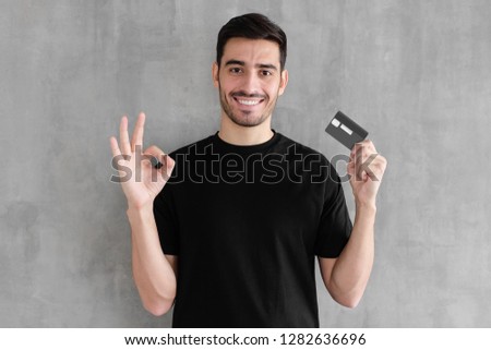 Portrait of young smiling man in black t-shirt, holding credit card and showing okay sign, standing against gray textured wall