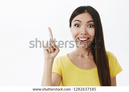 Energized young creative female genius with broad white smile raising index finger in eureka gesture adding suggesting having perfect idea or plan to share with team against gray background