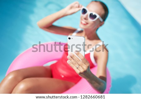 Vacation and technology. Colorful portrait of pretty young woman taking selfie on smartphone at swimming pool.