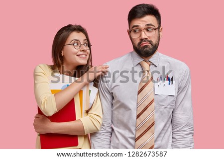 Horizontal shot of cheerful lady carries papers asks question in male classmate who has puzzled expression, cant give advice, isolated over pink background. Two partners work together with documents