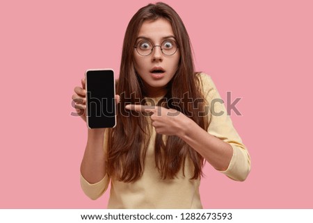 Headshot of surprised beautiful Caucasian woman has scared expression, points at mobile phone, demonstrates empty screen of gadget, isolated over pink background. People and technology concept