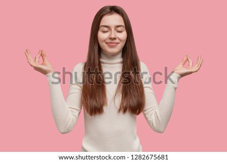 People and meditation concept. Relaxed peaceful young woman holds hands in mudra sign, keeps eyes closed, relaxes after work in office, wears casual white jumper, isolated over pink studio wall