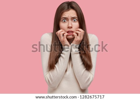 Nervous beautiful woman has worried look, feels puzzled and stressed before examination session, keeps hands near mouth, dressed in white jumper, being under pressure, models over pink studio wall