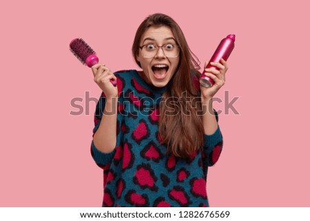 Photo of surprised European woman keeps mouth widely opened with positive expression, holds spray and hairbrush, has overjoyed look at mirror, cant believe in her new image, isolated on pink