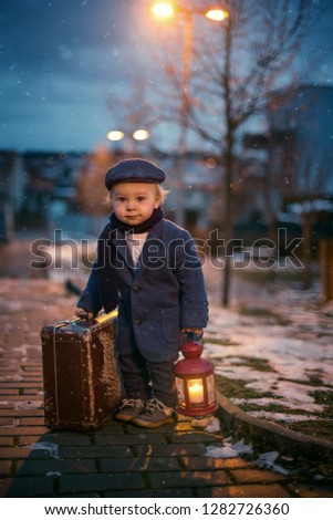 Toddler boy, standing on stairs, holding lantern and old suitcase, street view of Prague behind him, snowy evening