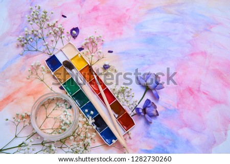 Composition with watercolor paints, brushes, a jar of water, flowers, flower petals, located in the figure with a watercolor background