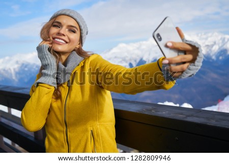 Smiling young woman in white coat and fur hat taking selfie outdoors 