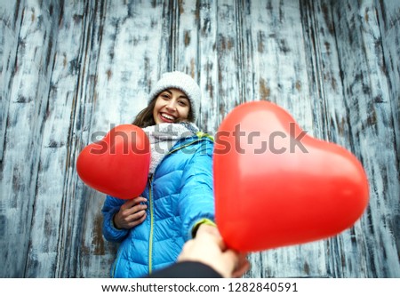 horizontal image of a happy smiling young woman is standing outdoors with a heart shaped red balloons on the old wooden door backround. woman is accepting a balloon from someone