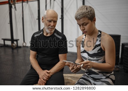 Picture of beautiful serious young European personal coach holding clipboard with workout plan before training with her elderly senior male client at fitness center, discussing exercises technique