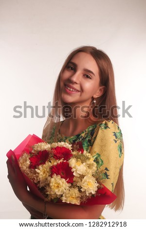 The portrait of smiling young blonde brunette lady with flowers isolated over white wall background wearing colorful bright dress looking camera