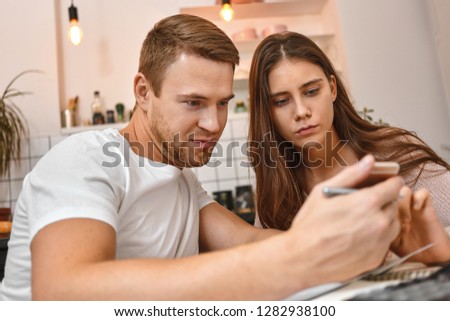 Modern gadgets and people. Picture of young couple having coffee in kitchen, using mobile phone reading online bank notification on unpaid mortage, looking at screen with worried facial expressions