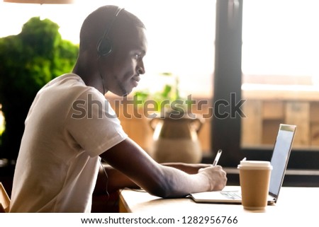 African student guy sitting at table studying preparing for university exam using headphones and notebook holding pen make some notes on textbook black man e-learning improve knowledge use application Royalty-Free Stock Photo #1282956766