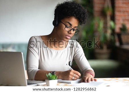 Serious african female smart student sitting at table wearing headphones listening online lecture writing notes on textbook. Self education and knowledge improvement using modern technologies concept Royalty-Free Stock Photo #1282956862