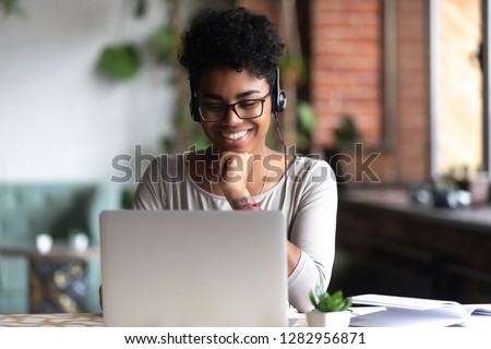 Black woman smart student girl sitting at table in university cafe alone wearing glasses looking at computer screen using headphones listening online lecture improve language skills having good mood Royalty-Free Stock Photo #1282956871