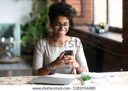 Cheerful african student black woman sitting at table studying using laptop reading a book, take a break holding mobile phone surfing internet received message from friend chatting about weekend plans Royalty-Free Stock Photo #1282956880