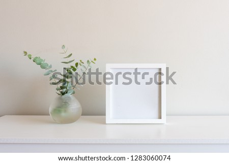 Eucalyptus leaves in small round glass vase next to blank square picture frame on white shelf against neutral wall background