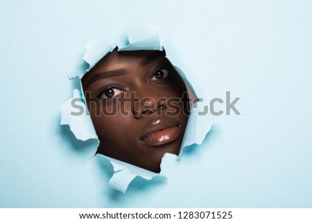 The face of a young beautiful girl with a bright make-up and puffy blue lips peers into a hole in blue paper. Fashion, beauty, make-up, cosmetics, hairstyle, beauty salon.