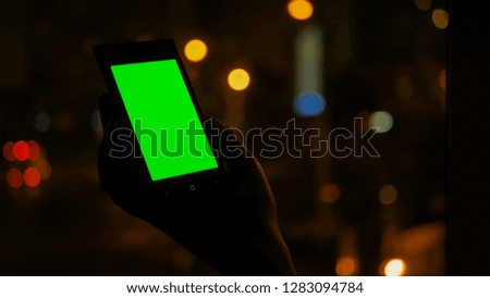 Woman looking at vertical smartphone with green screen at night. Blurred traffic bokeh light. Mock up and technology concept
