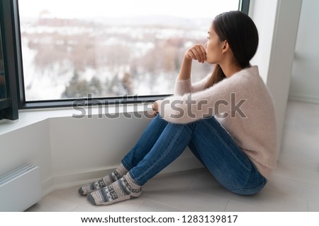 Depressed young girl feeling sad an lonely, anxious looking out the window in winter. Unhappy Asian woman alone, depression at home. Royalty-Free Stock Photo #1283139817
