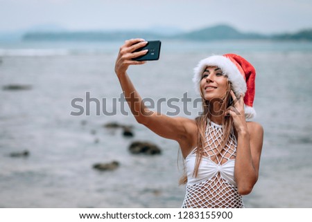 Summer beach vacation girl in santa hat taking fun mobile selfie photo with smartphone. Girl wearing white swimsuit posing for selfie. Phuket. Thailand. Vacation
