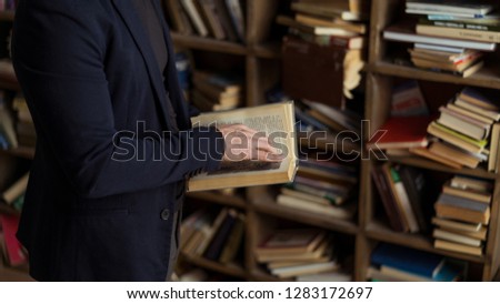 person in black navy suit holding book in his hands, shelves with books on background