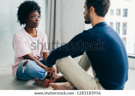 Young African woman and Caucasian man sitting near the window, staring at each other meditating together, free their minds from thoughts and worries.