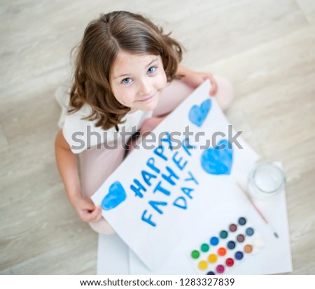 Little fun caucasian girl sitting on floor at home. Looking at camera. Cute kid paints a blue heart  on a homemade greeting card. Father Day. Traditional play concept. Arts and crafts. Top view