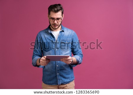Happy attractive young man standing and using tablet over pink background