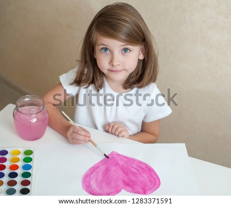 Child making homemade greeting card. A cute little girl paints a  pink heart on a homemade greeting card as a gift. Looking at the camera. Happy valentines day. Art and craft concept. Top view