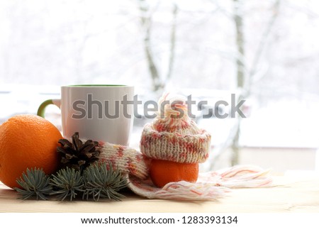 cup in a scarf and a mandarin in a hat decorated with a fir branch and a cone against the window. morning warming drink in winter