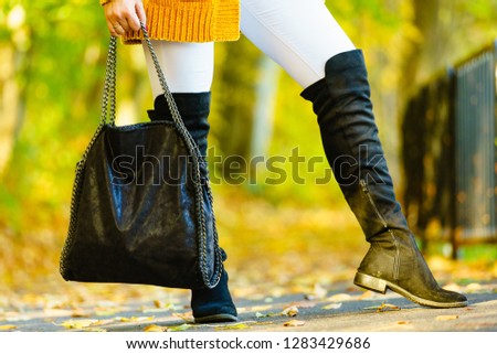 Woman presenting black suede high knee black boots holding big hand bag. Autumnal fashion, autumn season styled outfits. Female having a walk in park
