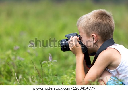 Profile close-up portrait of young blond cute handsome child boy with camera taking pictures outdoors on bright sunny spring or summer day on blurred light green grassy copy space background.