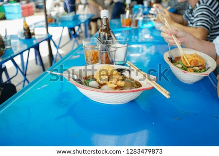 Closeup picture of spicy noodle with soup in a ceramic bowl with blurred people eating noodle in background. Delicious street food concept
