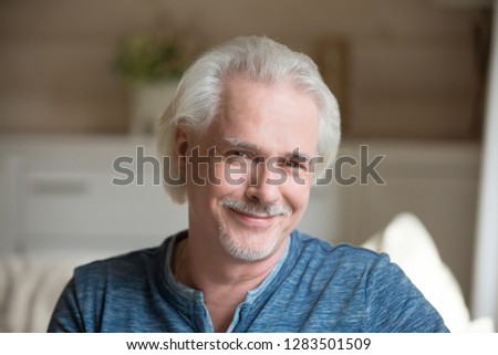 Head shot portrait of single handsome male sitting alone on couch in living room at country house in casual clothes smiling posing looking at camera. Successful healthy retired man full life concept